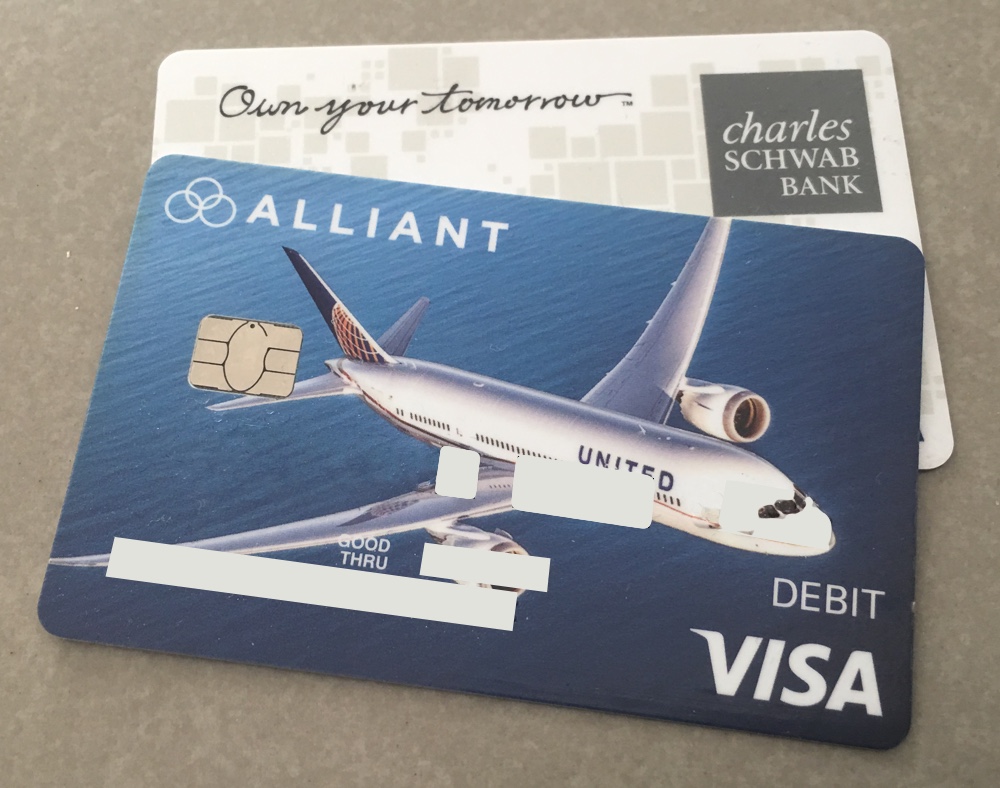 A photo of two debit cards, one from Charles Schwab Bank that reads "Own your tomorrow" superimposed over a map of the United States, and one from Alliant Credit Union featuring a United Airlines 787 airplane.