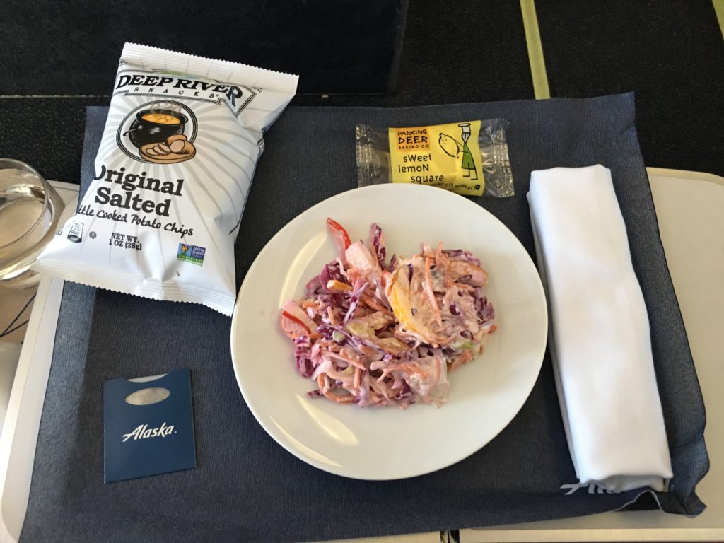 A first class meal try with a medium-size plate of coleslaw, a bag of potato chips, a lemon cookie bar, silverware wrapped in a white napkin, and a small packet of salt and pepper with an Alaska Airlines logo.