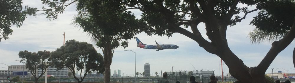 A WestJet Airlines plane with a Disney-themed paint job lands at LAX airport. There are large trees in the foreground that frame the airplane. Several buildings, a stoplight, streetlights, and a control tower are visible in the distance.