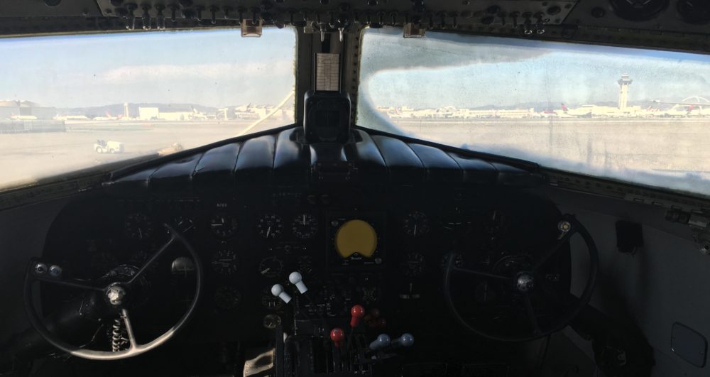 A photo of the controls and windows from the cockpit of a DC-3 aircraft. Part of the LAX tarmac and a control tower are visible through the windows.