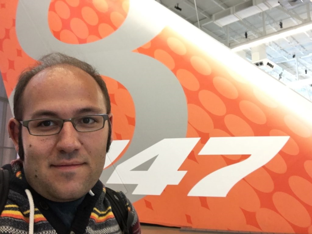 The author (wearing a hoodie with yellow, orange, red, and grey stripes) standing in front of an airplane tail. The tail is orange with a grey numeral 8 in the background and the numbers 747 in white in the foreground.