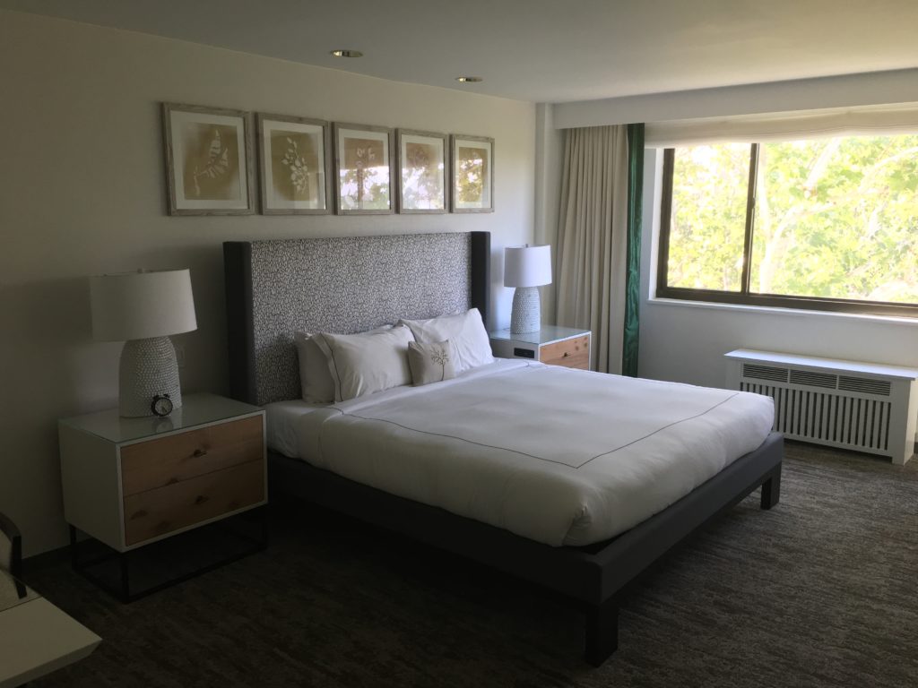 A photo of a king size bed with two side tables. Leaves are visible out the window.